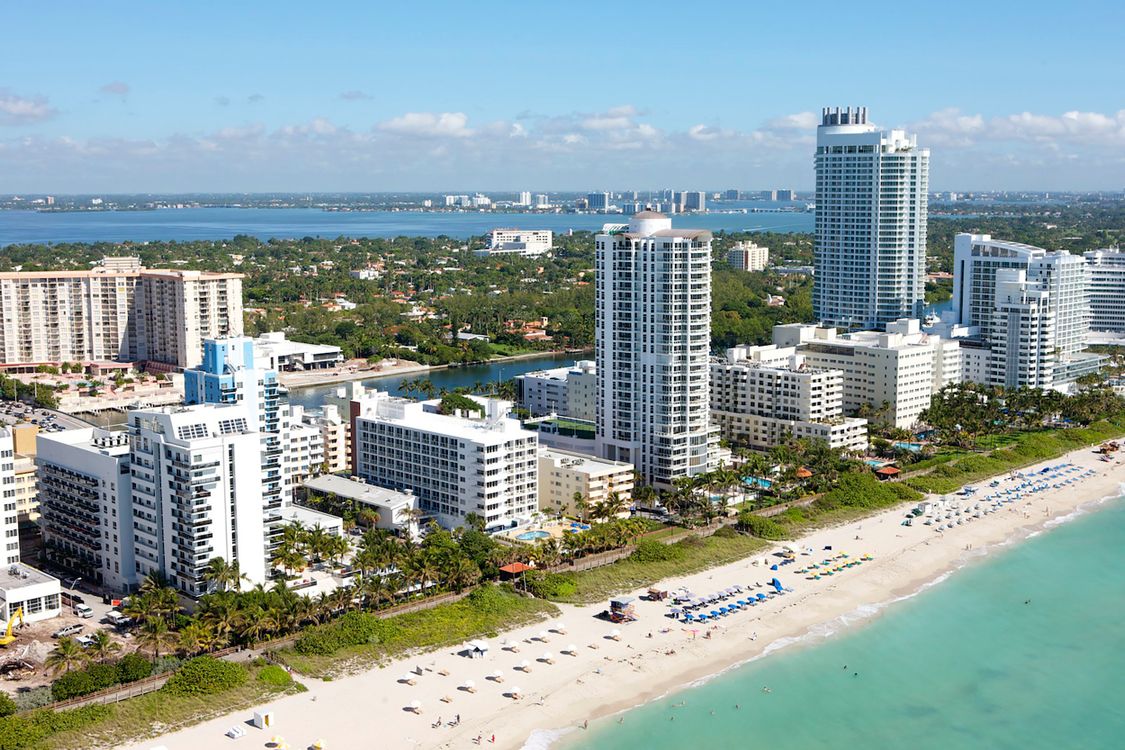 Aerial of Miami Beach oceanfront towers and beach with Biscayne Bay behind, showing the density buyers pay to escape
