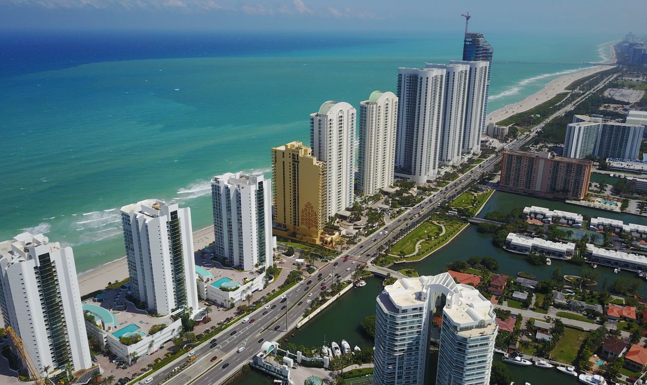 Aerial view of oceanfront condominium towers along Collins Avenue in Sunny Isles Beach, Florida, with a construction crane on a new tower, the type of pre-construction project marketed through an off-site sales gallery