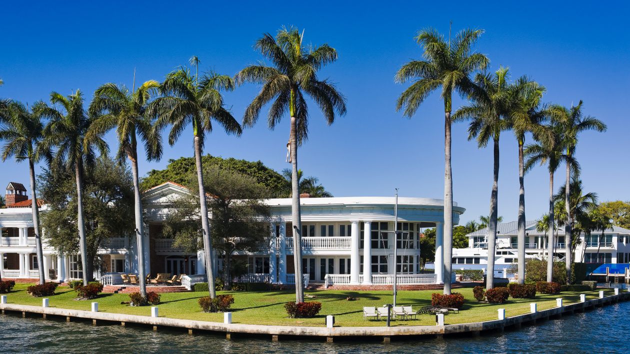 Colonnaded white waterfront estate framed by tall royal palms along a South Florida canal under a clear sky