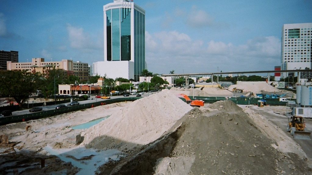 A graded Downtown Miami construction site with earthmoving equipment, the Metromover guideway and a tower behind it